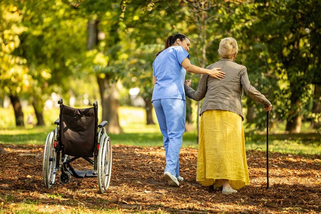 Two older adults and a caregiver talk together in a sunny common room with comfortable seating and large windows.