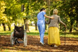 Two older adults and a caregiver talk together in a sunny common room with comfortable seating and large windows.